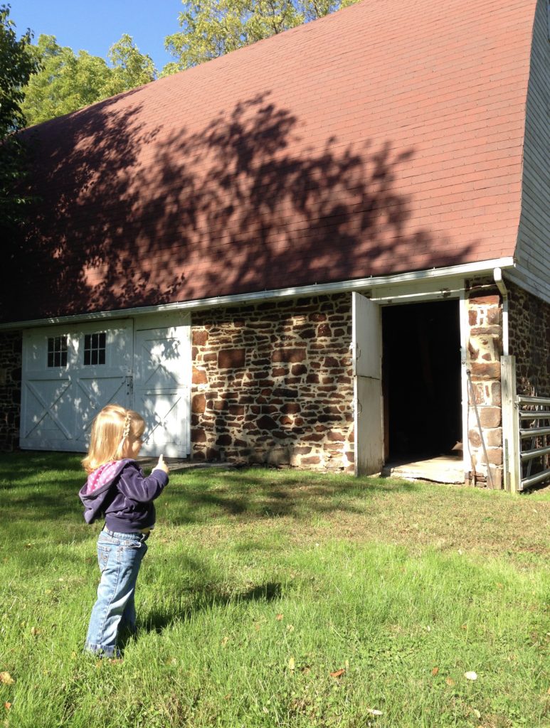 Old limestone barn with red roof and old barn doors. Blond toddler girl standing in front. 