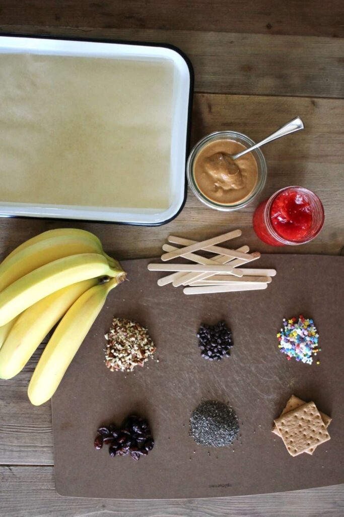 an enamel tray with wax paper, a bunch of bananas, popsicle sticks, peanut butter and jelly, and a variety of different toppings.