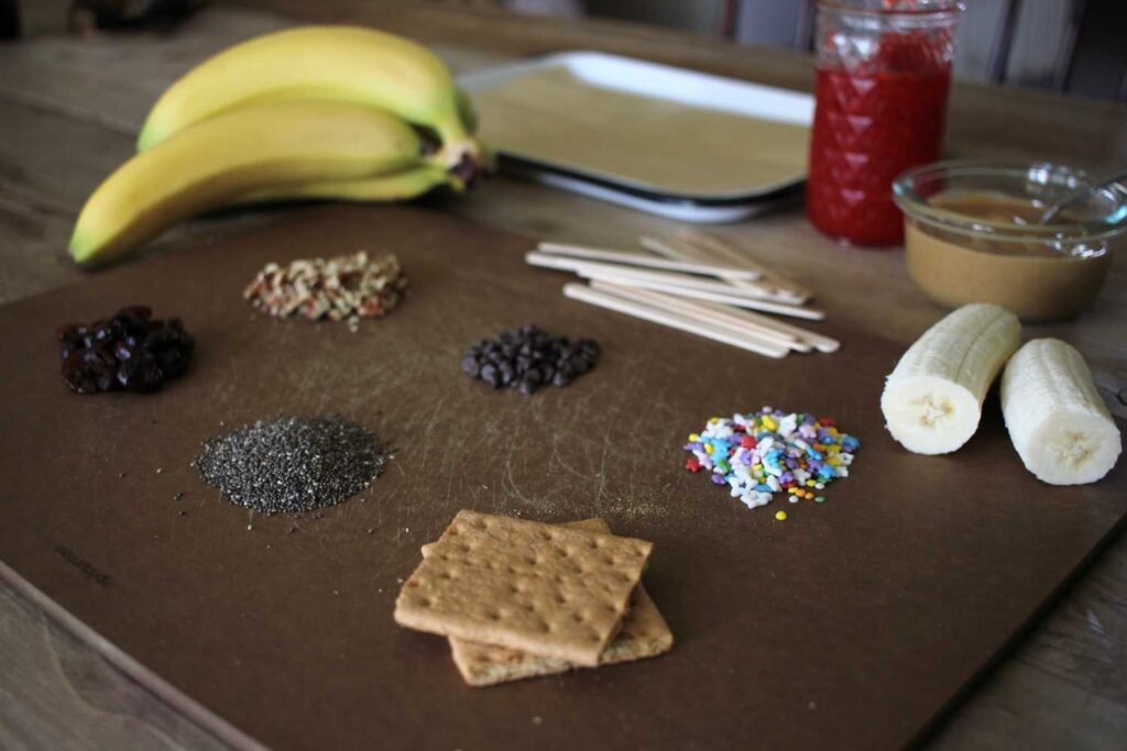 ingredients to make Banana Peanut Butter Popsicles; cutting board with different topping layer out a bunch of bananas and peanut butter and jelly 