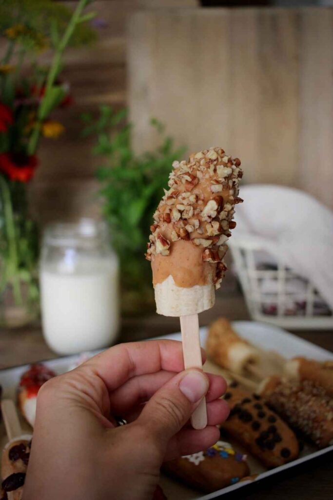 close up of a woman holding frozen banana on a popsicle stick covered in peanut butter with chopped pecans 
