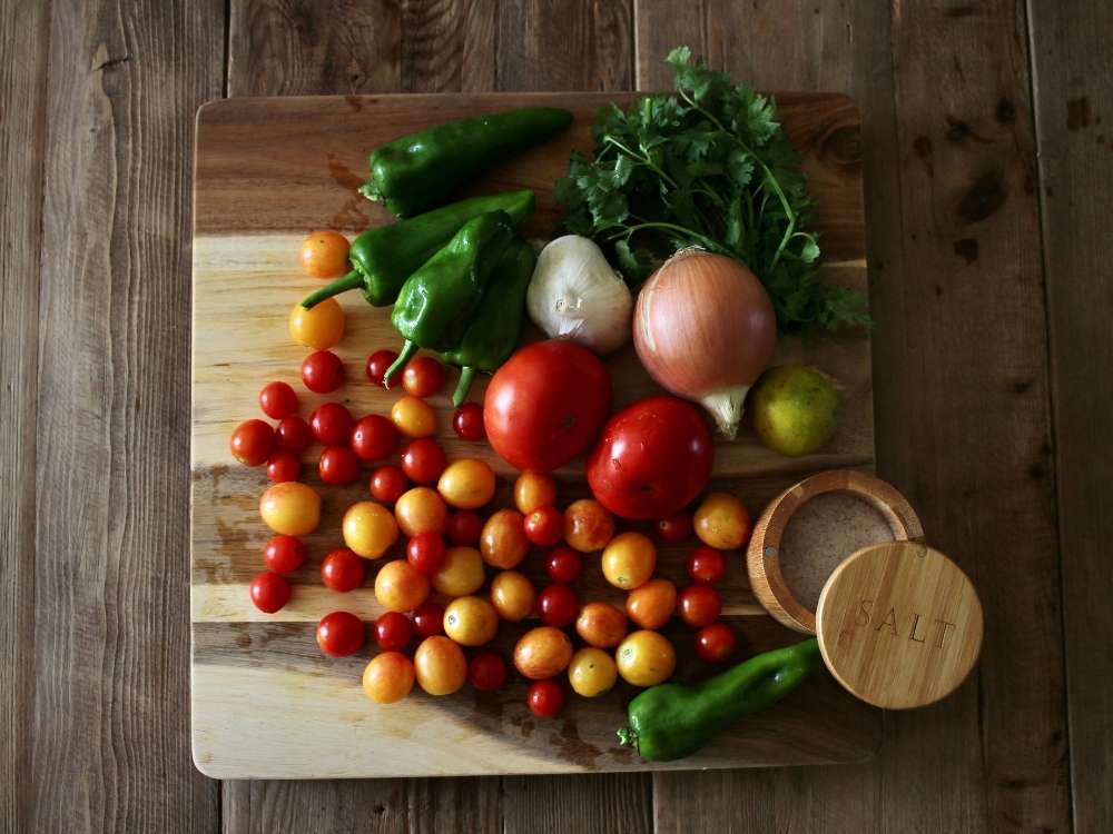 Ingredients for homemade salsa with tomatoes, peppers, onion, garlic, cilantro, lime, and salt laying on a large wood cutting board.