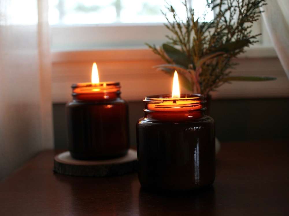 Two candles with large glowing flames sitting on a side table with a small bouquet of cedar and sage greens, underneath a window adorned with linen curtains.