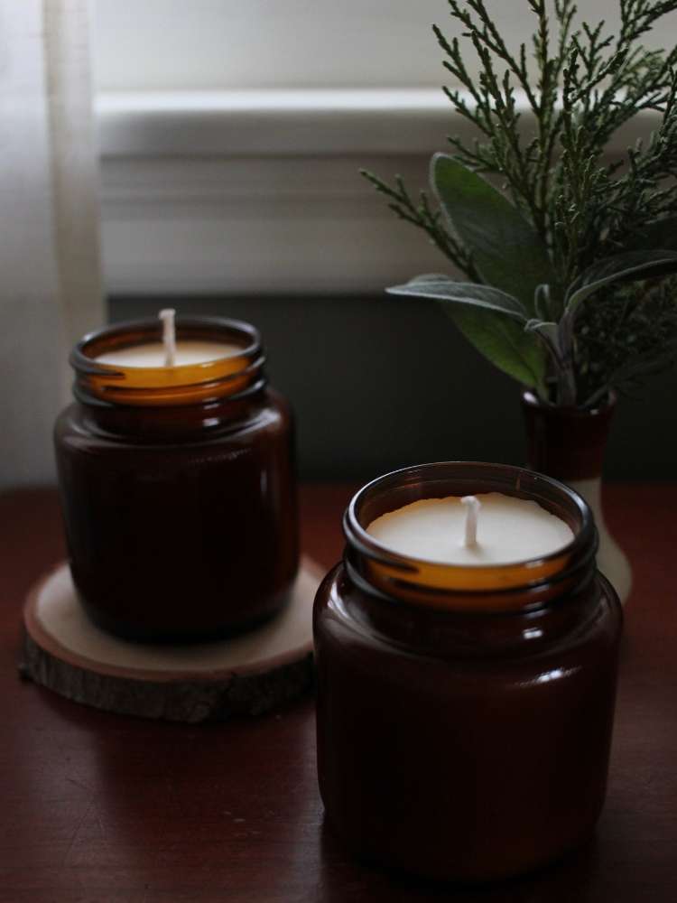 A close up of two homemade soy candles in recycled brown glass jars. In the background is a vase of green branches.