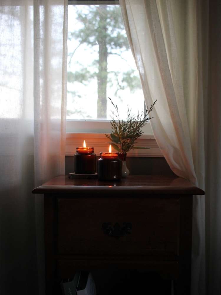 Underneath a large window with creamy linen curtains softly pulled aside, a pair of homemade soy candles in recycled brown glass jars are lit. 