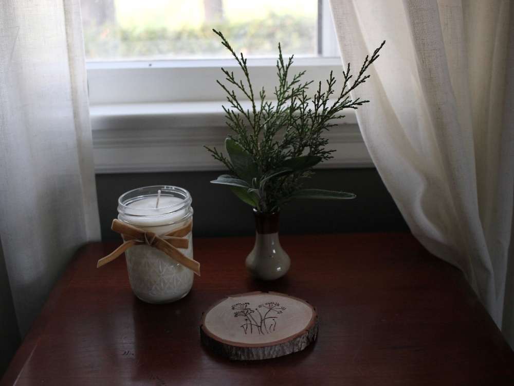 A dark moody picture with a new homemade soy candle adorned with a velvet bow. A small pottery vase of cedar and sage branches and a homemade wood coaster also sit on the side table. 