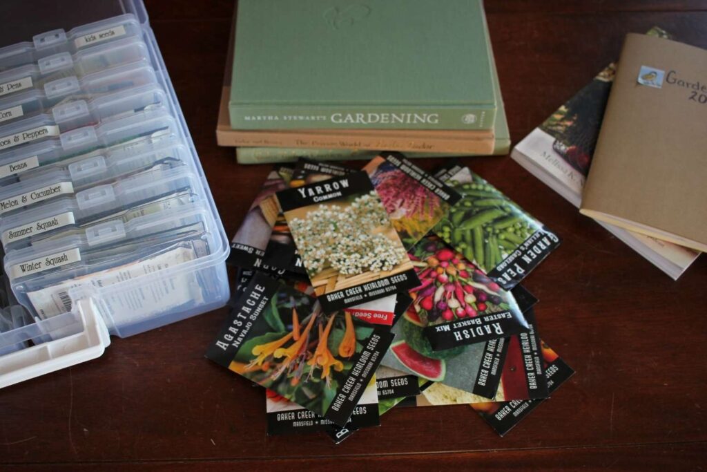 A bunch of seed packets laying on the table surrounded by gardening books and a seed organizing box.