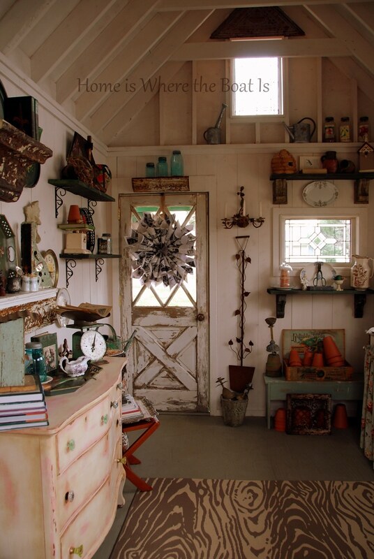 The interior of an a-frame garden shed with leaded stained glass windows a chippy paint door. Pots and other garden essentials and nicknacks are places about the room.