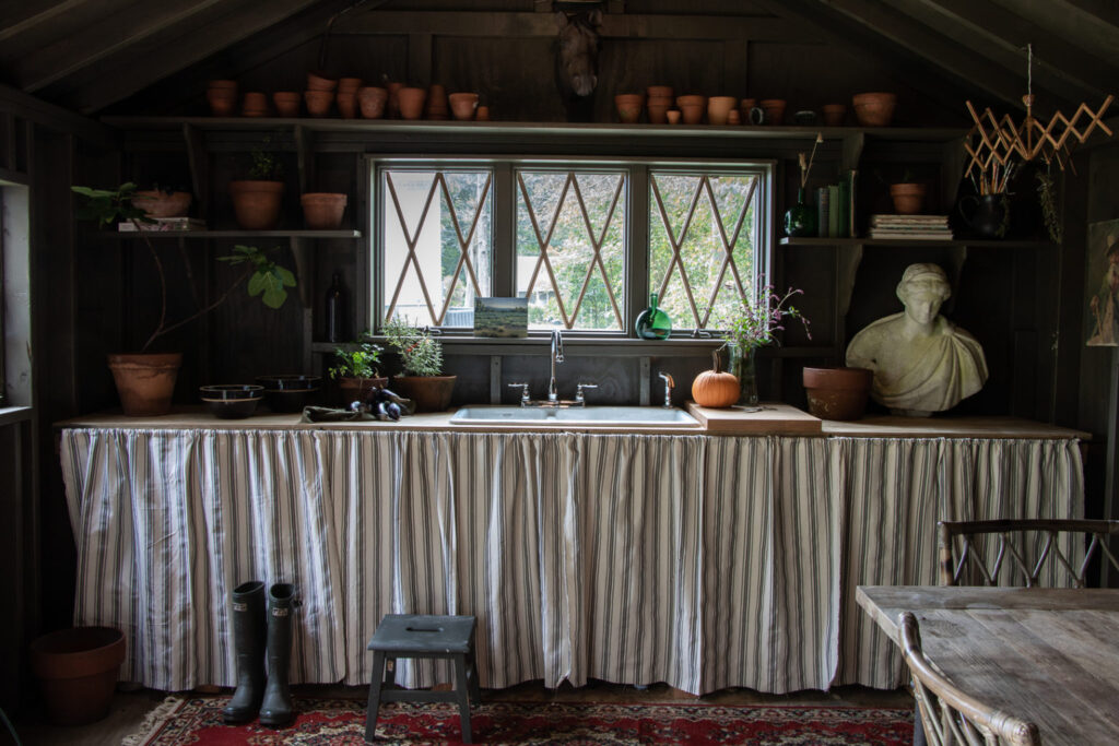 A dark green interior of a large garden shed. Pots line the shelves on the wall. A ticking striped skirt covers the front of the potting table.