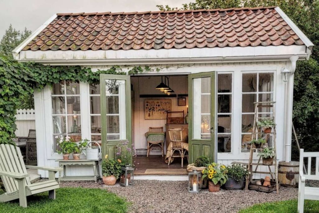 A orangery with green wood door and large paned glass windows. Vines grow up the side and aesthetically placed flower pots and garden decor surround the open doors.