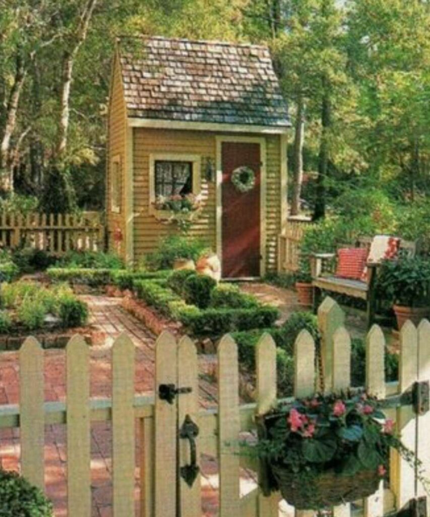 A tall and skinny garden shed with a red door and mustard siding place in the corner of a lovely manicured garden.