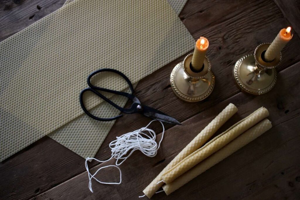 Rolled beeswax candles laying on a old wood table with two sheets of stamped honeycomb beeswax sheet laying beside. Two lit beeswax candles burn in brass candlesticks holder.