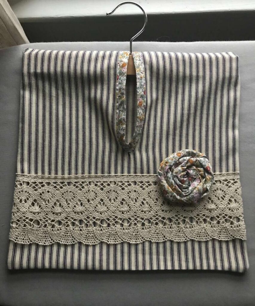 A simple vintage style clothespin bag with ticking striped blue fabric, crocheted lace at the bottom, and a fabric rose made from floral fabric.