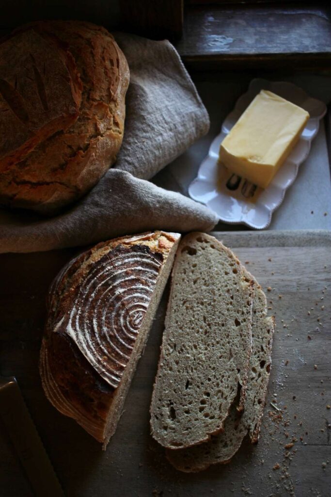 A loaf of fresh sourdough bread sliced with another loaf in the background and a dish a butter.