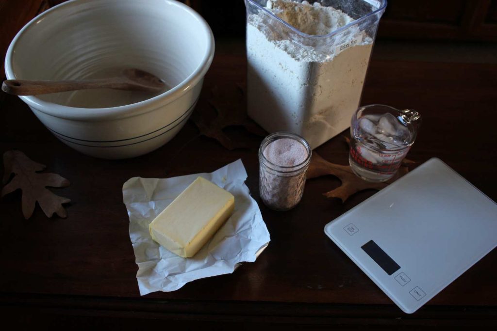 Ingredients for pie crust. A large ceramic cream colored bowl with two blue stripes with a wooden spoon inside. A tub of einkorn flour, a jar of pink salt, a measuring cup of ice water, a large stick of butter and a white scale. Several fall leaves on the table. 