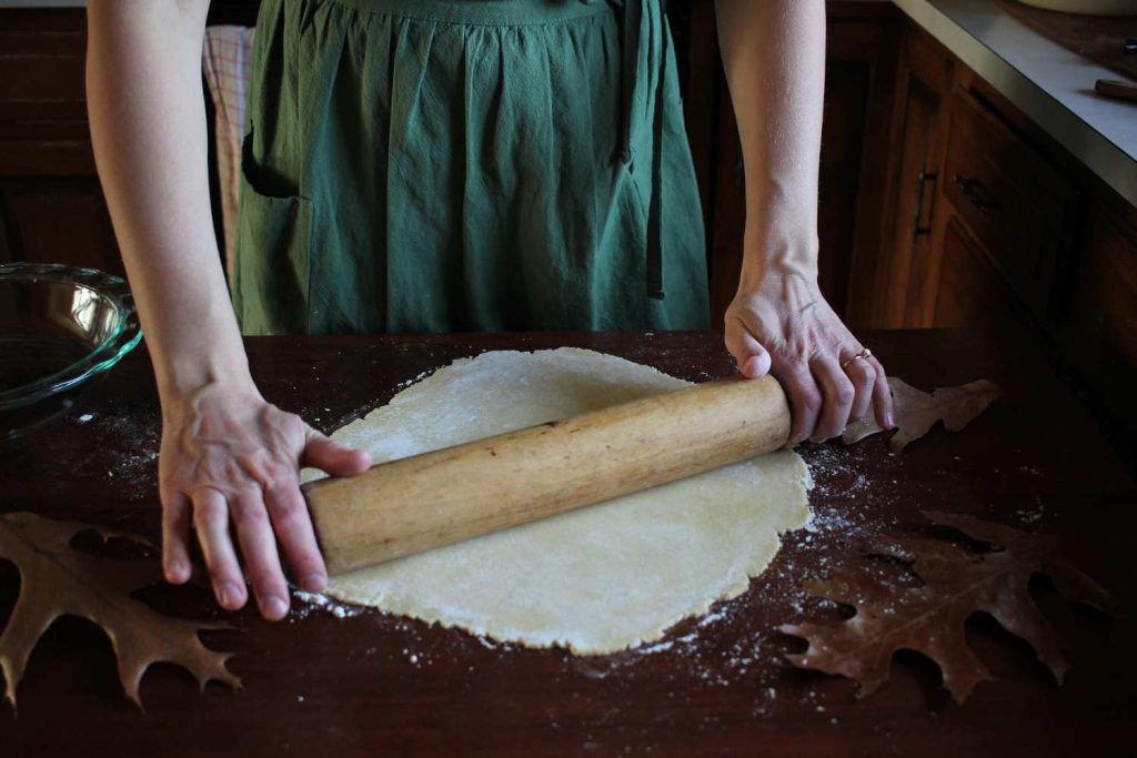 Close up of woman rolling out pie dough with a wooden rolling pin. Large brown fall oak leaves on the table. 