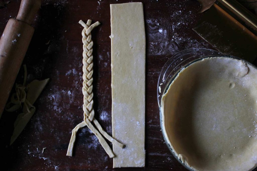 Pie crust placed into pie plate with extra pie dough rolled and cut into a large rectangular and three long thin strips of pie crust braided and a wooden rolling pin.