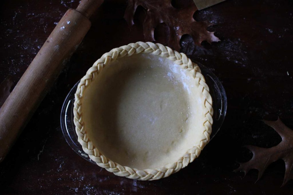 A close up of a homemade pie crust with a braided pie crust and rolling pie and maple leaves on the table surrounding the pie curst.
