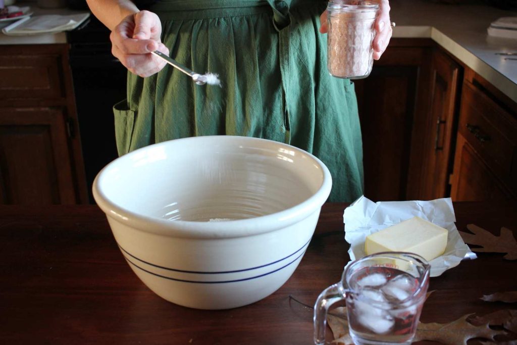 Woman with a green apron sprinkling pink salt into a large white ceramic bowl. Butter and ice water on the table. 