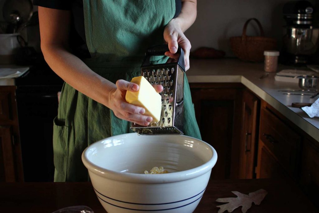 Woman's hand grating butter with a box grater into a large white ceramic bowl. 