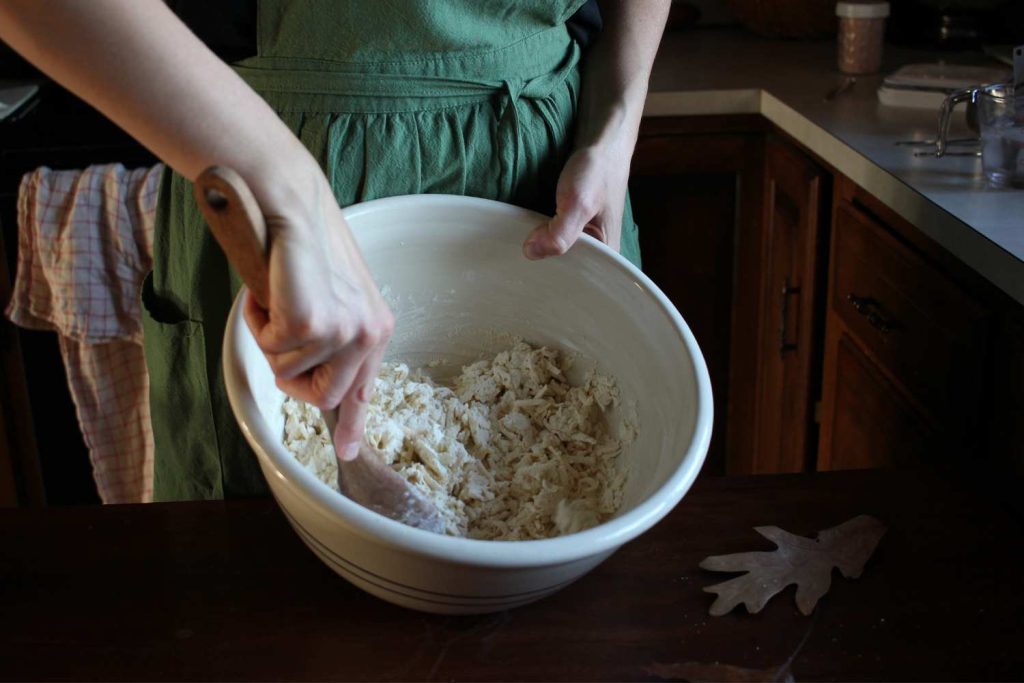 Woman's hands stirring flour, salt, grated butter in a large ceramic bowl.