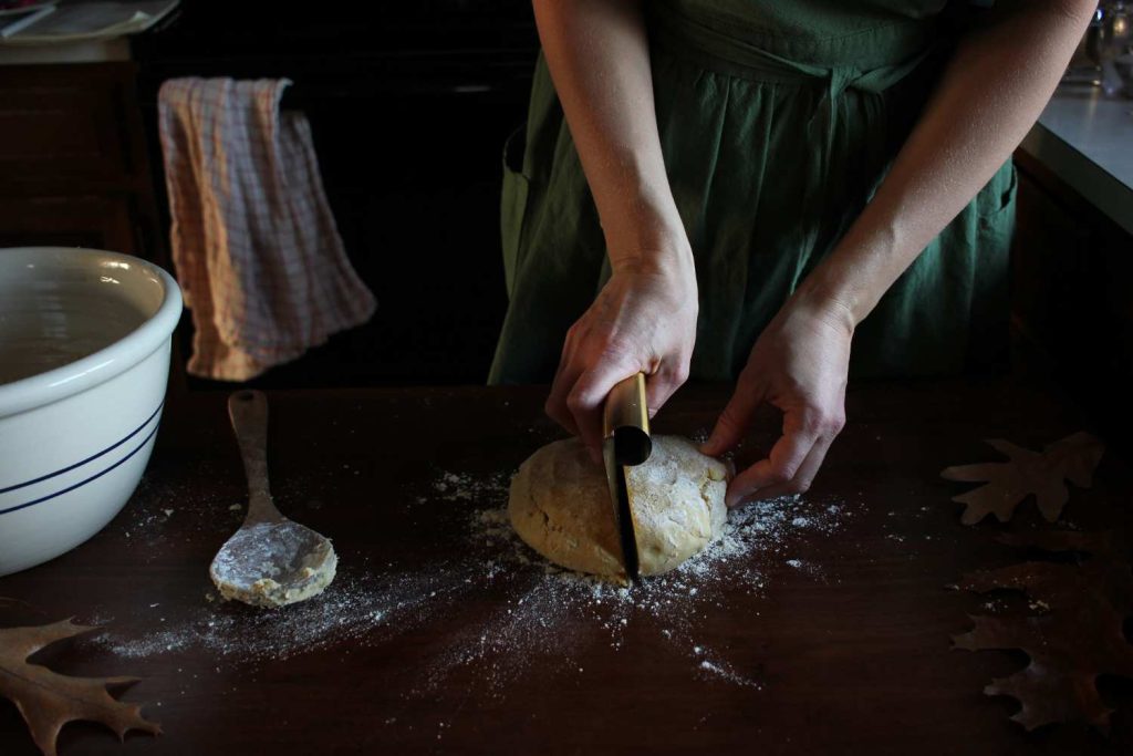 Woman's hands cutting formed pie dough into two pieces. Wooden spoon on the table with fall leaves and white ceramic bowl.