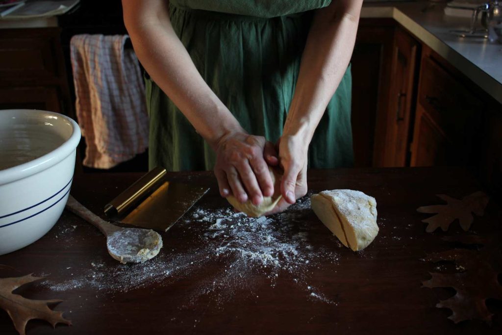 Woman's hands forming pie dough into a round flat circle. On the work table is more pie dough, a wooden spoon and bench scraper, large pottery bowl and several fall leaves.
