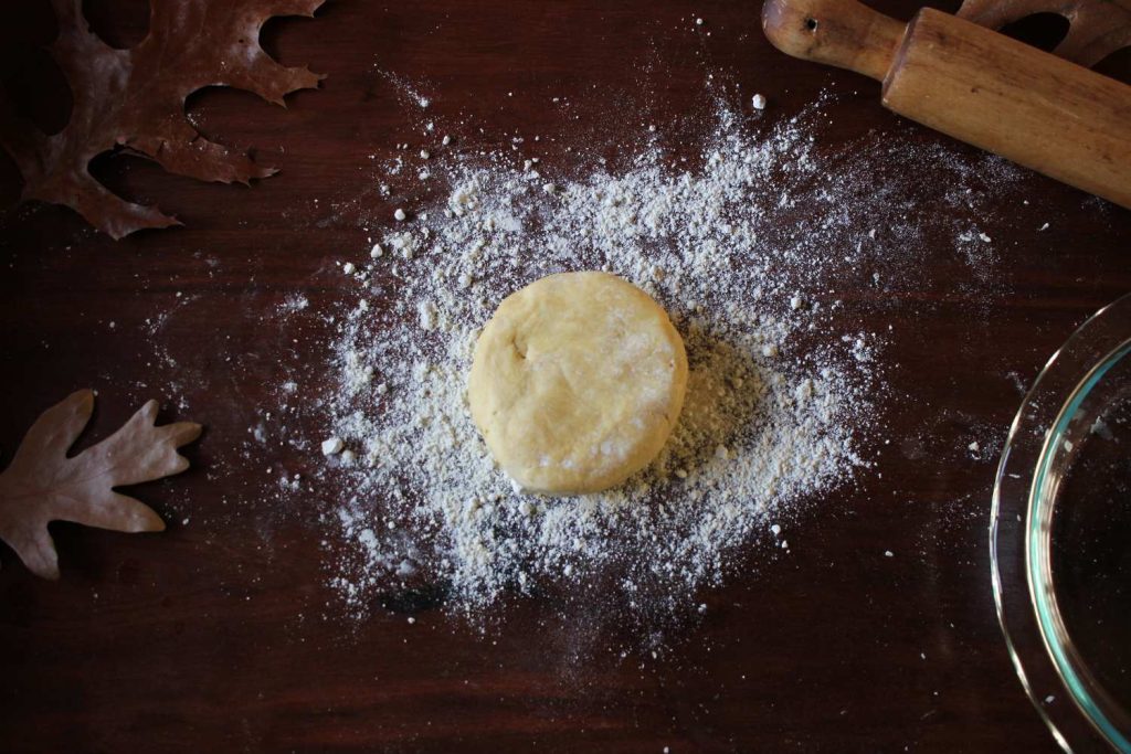 Close up of round circle of pie dough with flour sprinkled on table and a rolling pin and pie plate on the right side and fall leaves on the left.