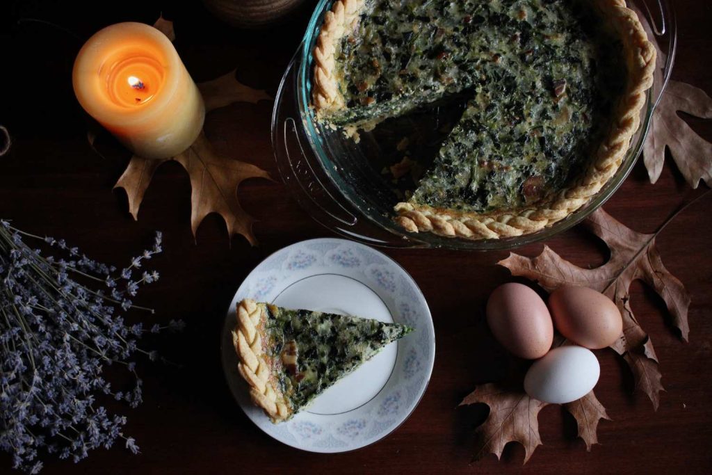 Bacon and spinach quiche with a slice of quiche on a white china plate with a light blue design on the edges. One the table around the quiche is a lit beeswax candle dried maple leaves, three brown and white eggs and a bouquet of dried lavender.