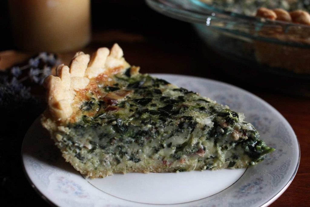 A close up photo of a slice of bacon and spinach quiche with a braided crust on a small china plate.