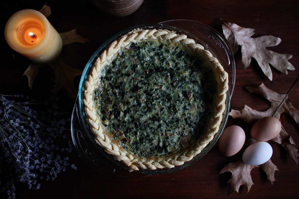 Bacon and spinach quiche with a braided pie crust on the table surrounded by a lit beeswax candle dried maple leaves, three brown and white eggs and a bouquet of dried lavender.