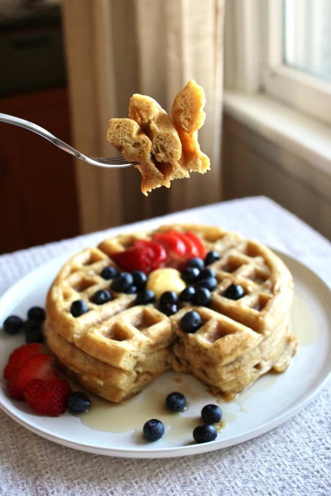 A fork with three bites of sourdough waffle on it and below and blurred a plate of sourdough waffles and fresh fruit on top.