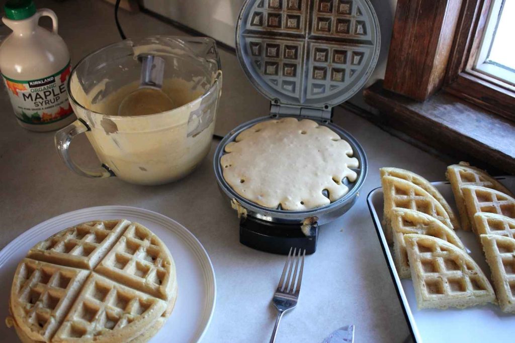 Waffle batter poured onto a waffle iron with a bowl of sourdough waffles to the left and a plate and tray of sourdough waffle below.