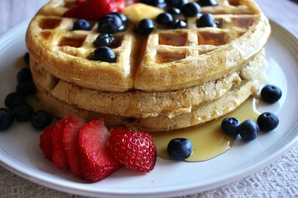 A stack of waffles with melted butter, strawberries and blueberries and maple syrup drizzling down the sides.